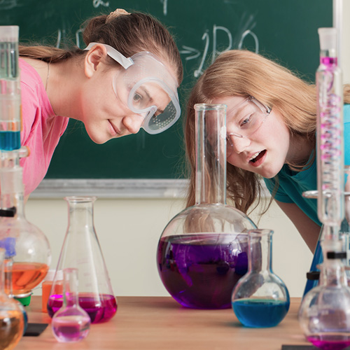 Two students wearing lab goggles staring at a beaker full of purple liquid.