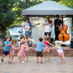 Children dancing around in a circle while a band performs behind them.