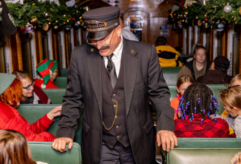 Man dressed in train conductors outfit walks up aisle on old train car. Children and families in pajamas are seated in the rows.